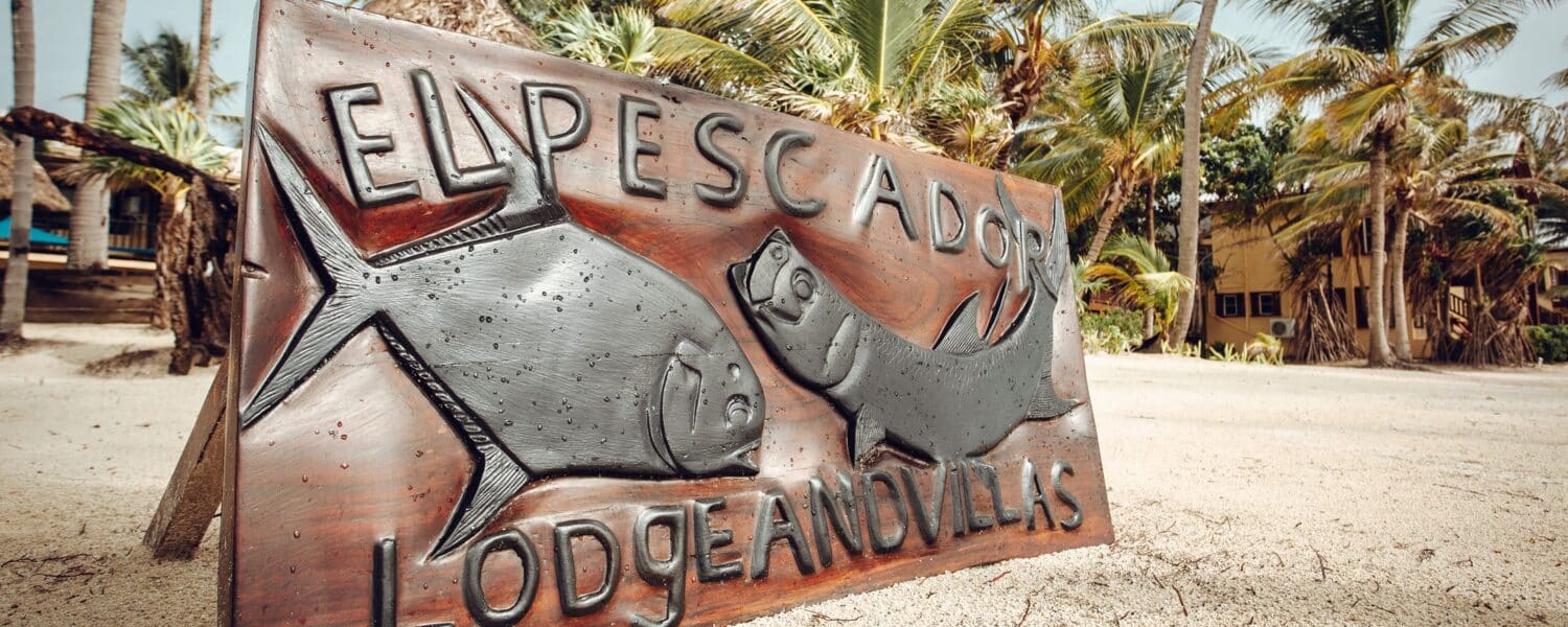 El Pescador Lodge beachfront sign surrounded by tropical palms and sandy shores.