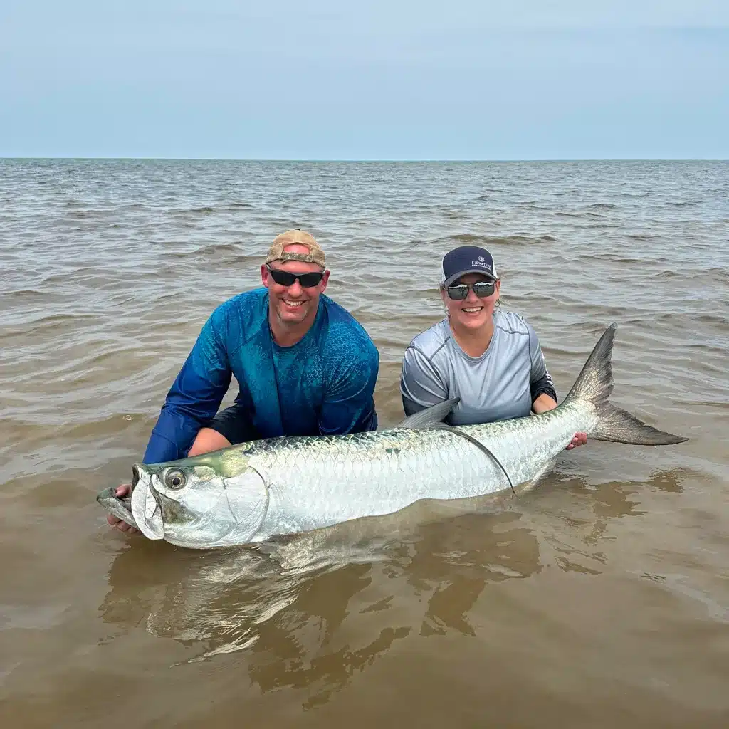 Two anglers proudly display their catch: a giant tarpon in shallow waters.