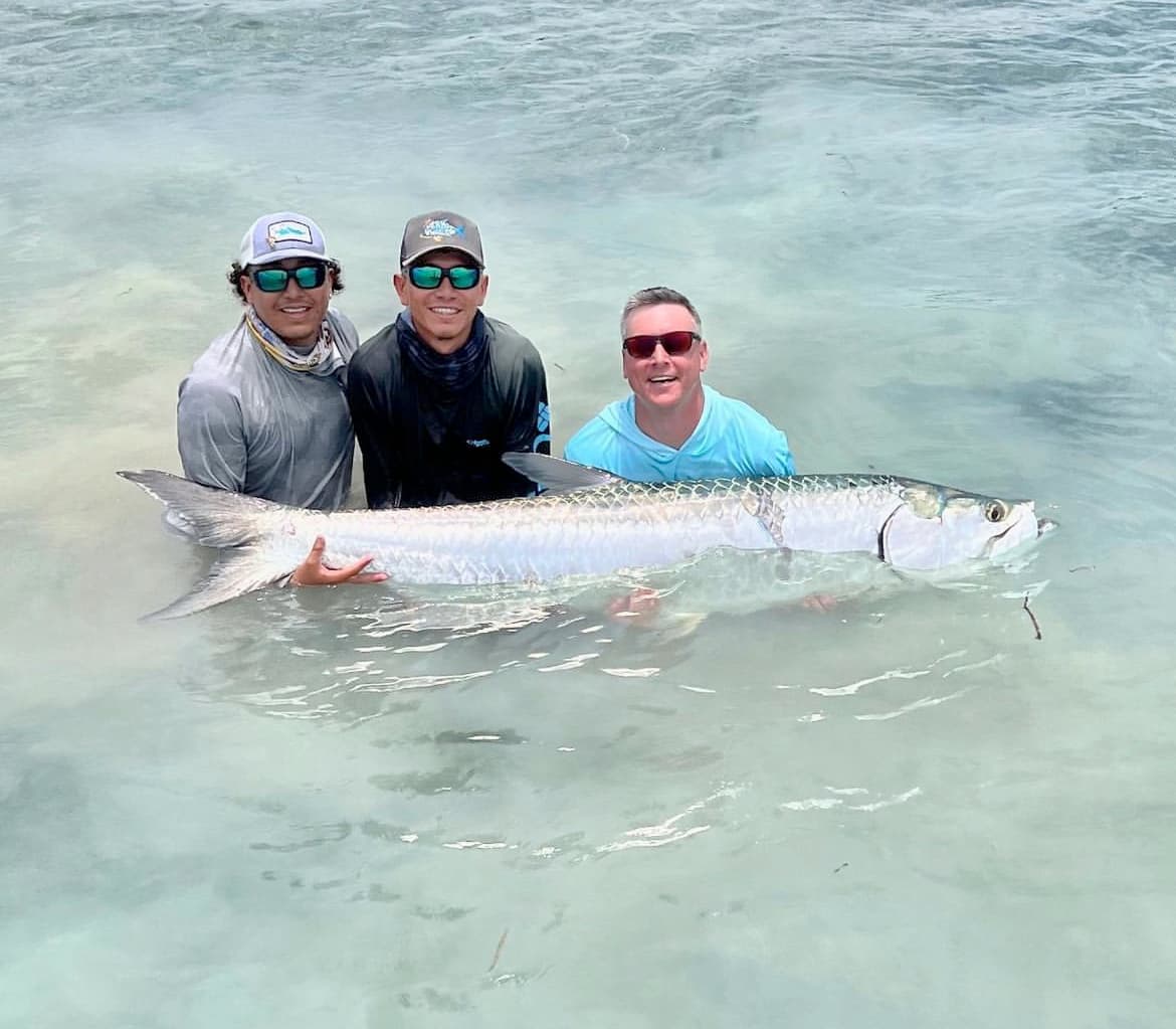 Three anglers proudly display their impressive tarpon catch in crystal-clear water.